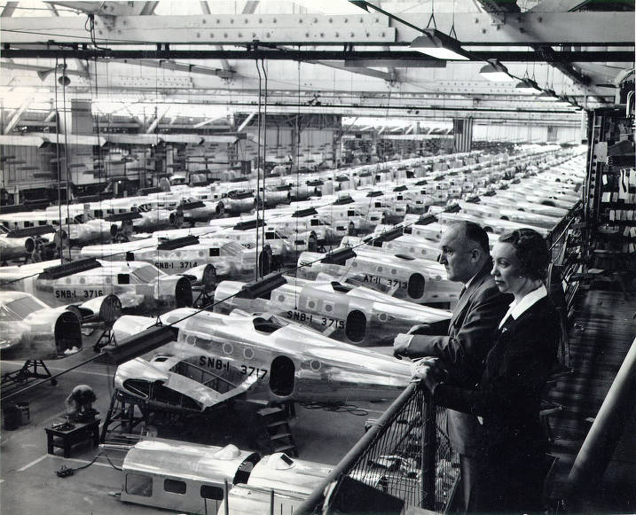 Insert image here: Elliptical wings in assembly jigs; fitters riveting skins while inspectors check with gauges and templates.